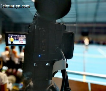 A camera on a tripod is prominently displayed with its viewfinder screen showing footage of an indoor sports event. The background is an indoor sports hall with a blue floor, and numerous spectators are faintly visible.