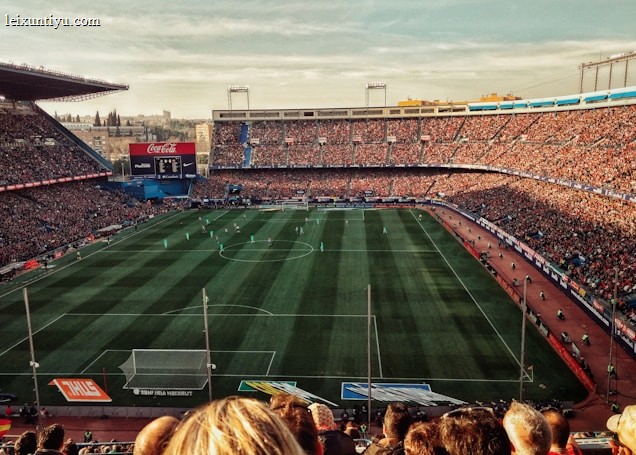 A large football stadium filled with spectators watching a match. The green pitch is lined with players in action, surrounded by packed stands. Banners and advertisements are visible around the stadium.