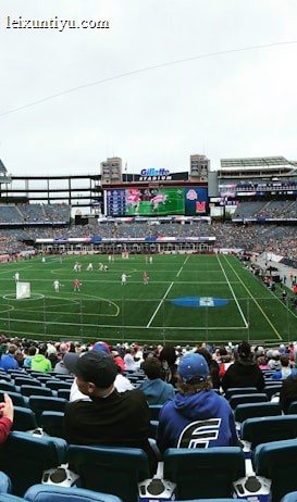 A large sports stadium filled with spectators. The field is green with white markings, and players in red and white uniforms are actively engaged in a game. The stands are packed with people wearing various colored clothing, and some sections of the crowd are more densely populated. Digital screens and advertisements are visible around the stadium, adding to the vibrant atmosphere.