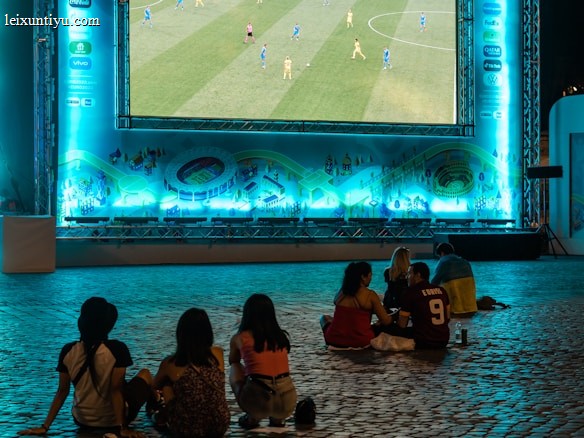 A large outdoor screen displays a soccer match at night. A small group of people sit on a cobblestone surface, watching the game. The screen is surrounded by a bright blue glow, creating a lively atmosphere.