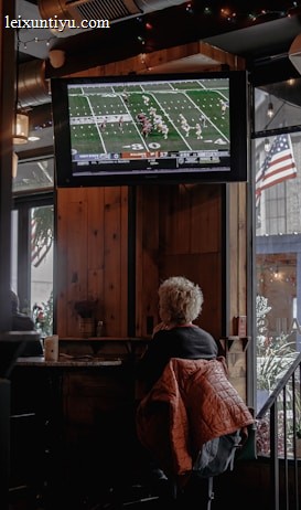 A person with short, light-colored hair is sitting in a cozy, wood-paneled interior of what appears to be a bar or cafe, watching a football game on a mounted TV screen. A warm-colored jacket is draped over their chair. The TV displays a live football match with a clear scoreboard and players on the field. The space is decorated with string lights, and an American flag is visible outside through a window next to some greenery.
