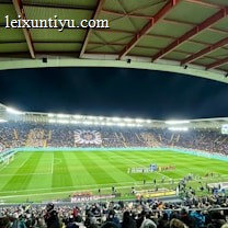 A large football stadium filled with spectators. The field is brightly lit, and two teams are positioned on the pitch. The stadium is covered, with a visible roof structure and stands filled with fans. Scoreboards are visible displaying game information.
