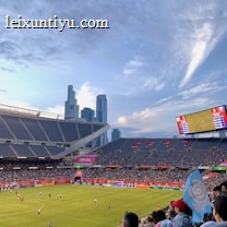 A sports stadium filled with spectators watching a soccer match. Players are on the field in action, and a large scoreboard displays information about the game. The stadium has blue seating and is surrounded by tall city buildings visible in the background against a partly cloudy sky. Fans are dressed casually, some waving flags and wearing team colors.