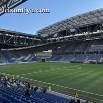 A modern sports stadium with a large, green soccer field and surrounding seating areas. Blue and yellow seats are visible, with some spectators scattered across the stands. The stadium is partially covered with a steel lattice roof, showing a clear blue sky. Several security personnel in high-visibility vests are present.