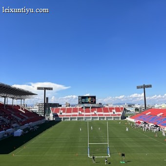 A large sports stadium with red and blue seating overlooks a green field with a goalpost at the center. A few people are scattered across the field, with some tents and a gathering of individuals on the right side. The sky is clear and blue with some clouds visible in the distance. A large screen or scoreboard is situated at the far end of the field.
